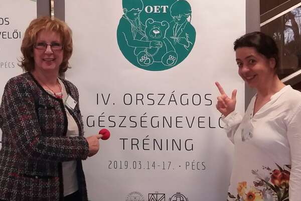 Két nő egy nagyméretű tábla mellett a kamerába néz
